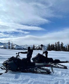 two people standing next to a snowmobile in the snow with mountains in the background