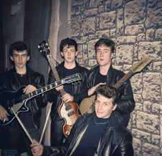 four young men are posing with their instruments in front of a brick wall, one is wearing a black leather jacket and the other has a guitar