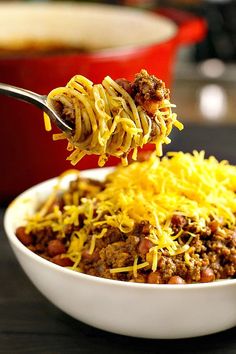 a spoon full of chili and cheese is being held over a bowl filled with spaghetti