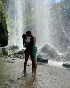 two people standing in front of a waterfall with their arms around each other and kissing