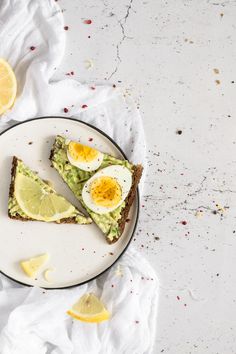 two slices of avocado toast on a white plate with lemon wedges next to it