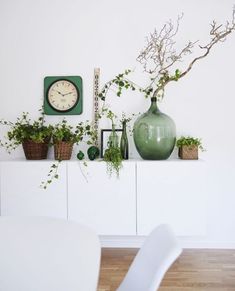 a green vase sitting on top of a white table next to a clock and plants