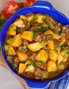 a blue bowl filled with meat and potatoes on top of a wooden cutting board next to tomatoes