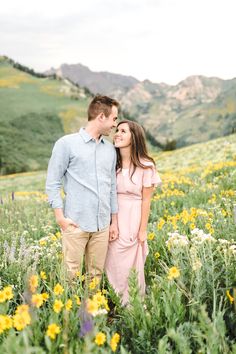 a man and woman standing in a field full of wildflowers with mountains in the background