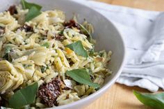 a white bowl filled with pasta and greens on top of a wooden table next to a napkin