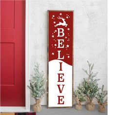 a red and white sign that says believe next to some christmas trees in front of a red door