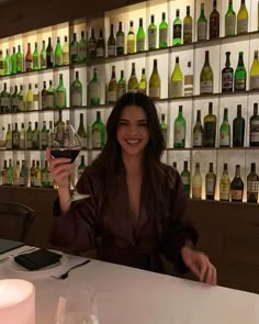 a woman sitting at a table holding up a wine glass in front of bottles on the wall