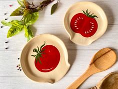 two bowls with tomatoes painted on them next to a wooden spoon and leafy plant