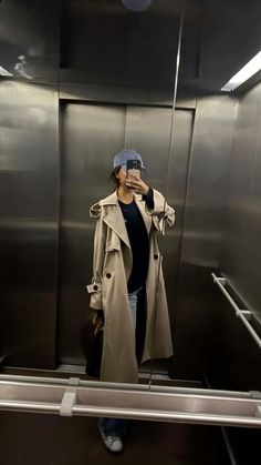 a woman taking a selfie in an elevator while wearing a trench coat and hat