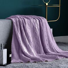 a purple blanket sitting on top of a gray couch next to a white vase and lamp