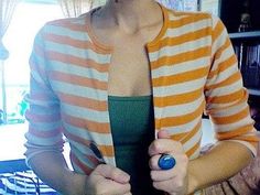 a woman in an orange and white striped cardigan holding a blue object while standing next to a bookshelf