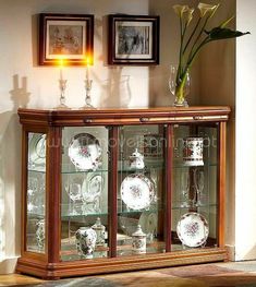 a glass cabinet with plates and vases on it in a room that has framed pictures