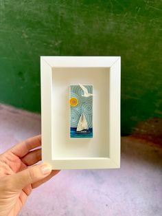 a hand holding up a small white frame with a sailboat in the ocean on it