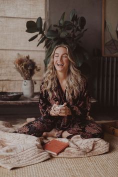 a woman sitting on the floor laughing and holding a cup in her hands while looking at something