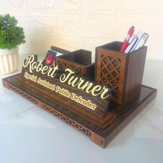 a wooden desk organizer with pens and pencils in it sitting on a table next to a potted plant