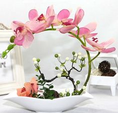 pink flowers are in a white bowl on a table next to framed pictures and other decorative items