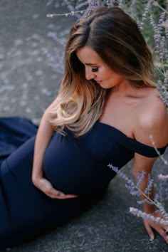 a pregnant woman sitting on the ground in front of some purple flowers wearing a blue dress