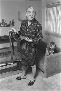 an old man in pajamas standing next to a dog on a couch and holding a book