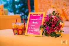 a table topped with a sign and flowers next to glasses filled with drinks on top of it