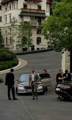 three men in suits standing next to two parked cars