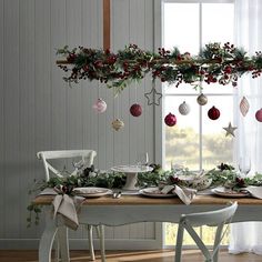 a dining room table decorated for christmas with greenery and ornaments hanging from the ceiling