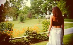 a woman in a long white skirt is walking through the grass with flowers behind her