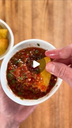 two hands holding a small bowl of chili with cheese on top and chips in the other hand