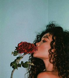 a woman with long curly hair is smelling a red rose