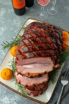 a large ham sitting on top of a white plate next to oranges and wine glasses