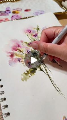 a hand holding a pen and drawing flowers on a piece of paper with watercolor pencils