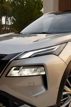 the front end of a silver car parked in front of a building with trees behind it