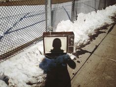 the shadow of a person standing next to a television on snow covered ground in front of a chain link fence