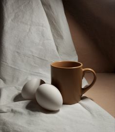 two eggs sitting next to a coffee cup on top of a white cloth covered table