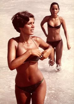 two women in bikinis are standing in the water at the edge of the beach