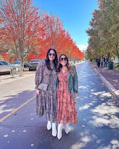 two women standing next to each other in front of trees with orange and red leaves