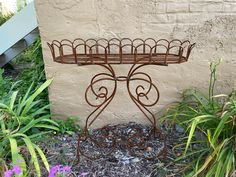 a wrought iron plant stand in front of a wall with flowers and plants around it