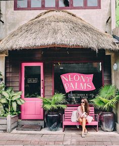 a woman sitting on a pink bench in front of a building with thatched roof