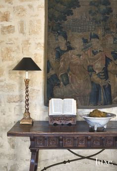 a table with an open book on it next to a lamp and a large tapestry