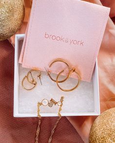 three pairs of earrings in a box on a pink surface with gold ornaments around it