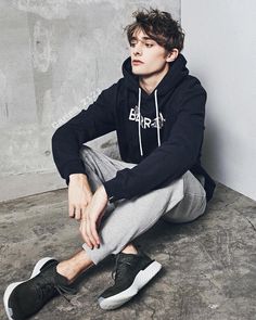 a young man sitting on the floor wearing a black hoodie and grey sweatpants