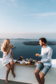 a man and woman sitting at a table with food in front of the ocean on a sunny day