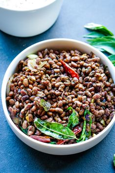 a white bowl filled with beans and greens next to a bowl of rice on a blue surface