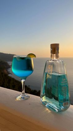 a blue drink sitting next to a bottle on top of a wooden table near the ocean