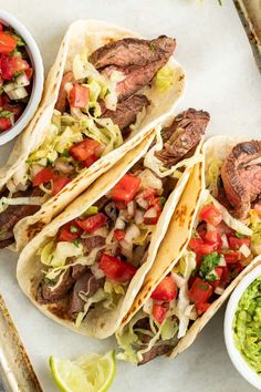 three steak tacos with guacamole, salsa and lime wedges on the side