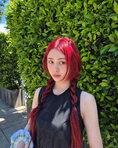 a woman with long red hair standing in front of a bush and looking at the camera