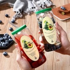 two people holding up jars filled with blueberries and orange juice on top of a wooden table