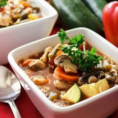 two bowls filled with soup and vegetables on top of a red cloth next to silver spoons