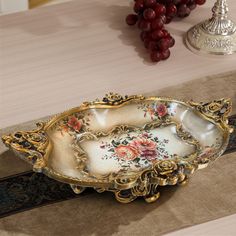 an ornately decorated serving dish sits on a table with grapes and other items in the background