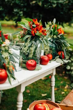 an outdoor table with apples and greenery on it, in front of some flowers