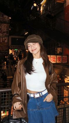 a woman standing next to a fence holding a black bag and wearing a brown hat